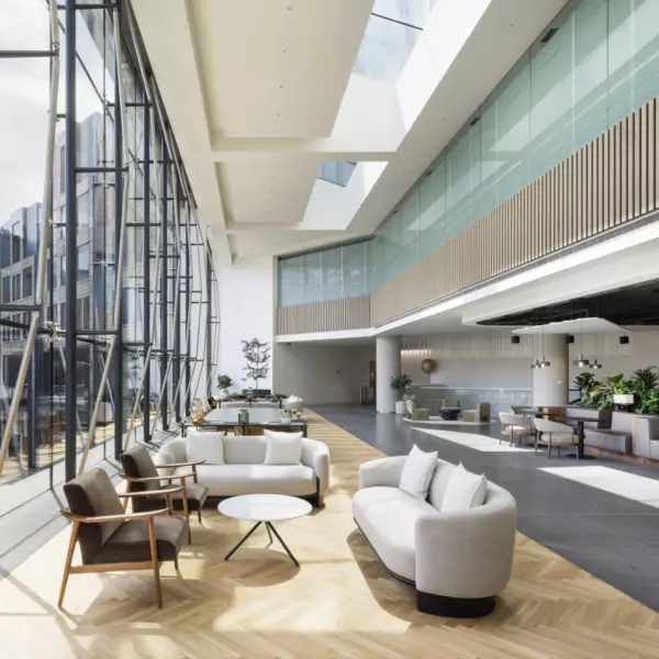 A spacious modern office interior with floor-to-ceiling windows on the left, flooding the space with natural light. Contemporary seating includes sofas and armchairs around small tables. Light wood and dark tile flooring define zones, while pendant lights, plants and glass panels with wooden slats enhance the sleek, welcoming design.