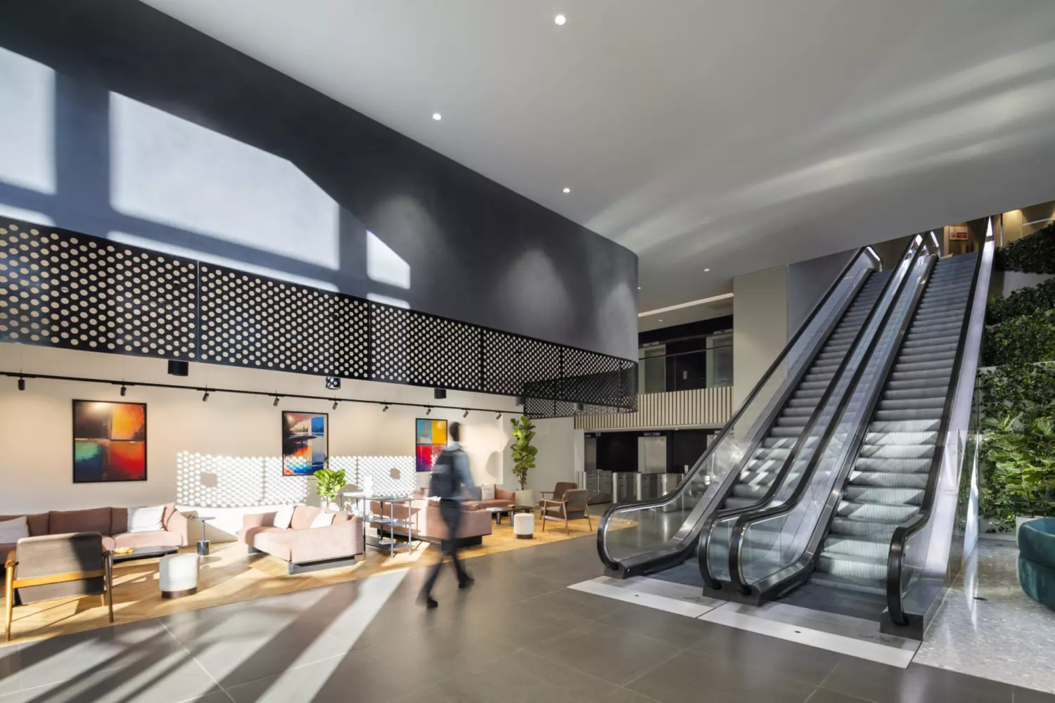 A modern indoor office space featuring a relaxed seating area with sofas, chairs and tables, accented by colourful wall art. An escalator on the right leads to an upper level, surrounded by greenery. Large windows fill the space with natural light, creating a bright, welcoming atmosphere.