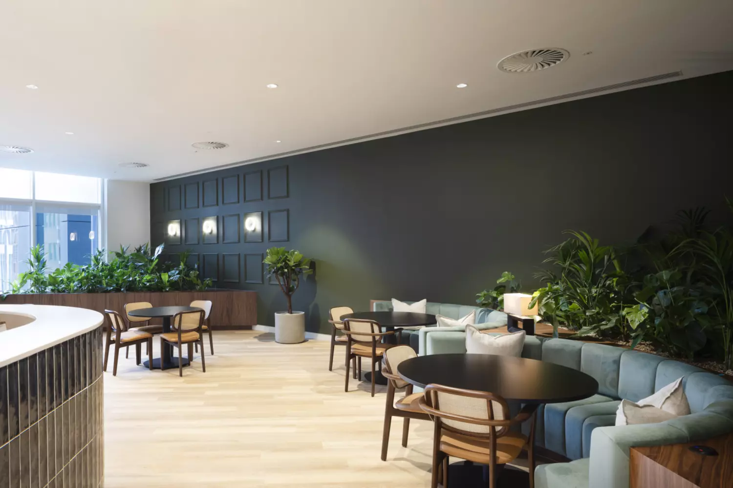 A modern lounge area with wooden flooring and varied seating including round tables with chairs and a long cushioned sofa. Green plants in pots and built-in planters add natural texture. The back wall features dark square panelling with integrated lighting, while large windows on the left bring in natural light.