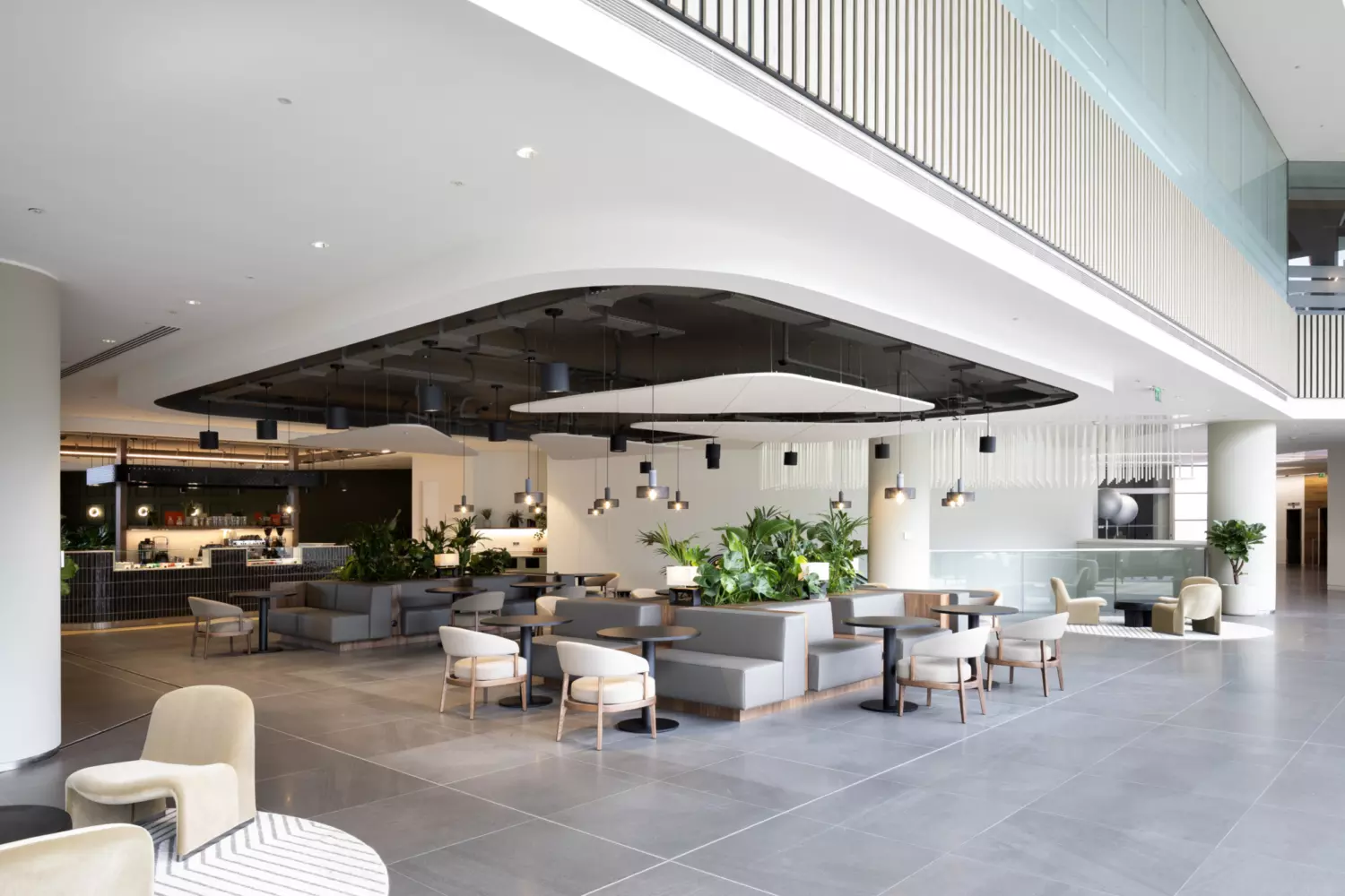 A spacious modern indoor area with sofas and chairs arranged around tables, illuminated by natural light from large windows. Hanging ceiling lights and greenery enhance the relaxed atmosphere. The tiled grey flooring leads to a café-style counter in the background, with additional seating visible further into the space.