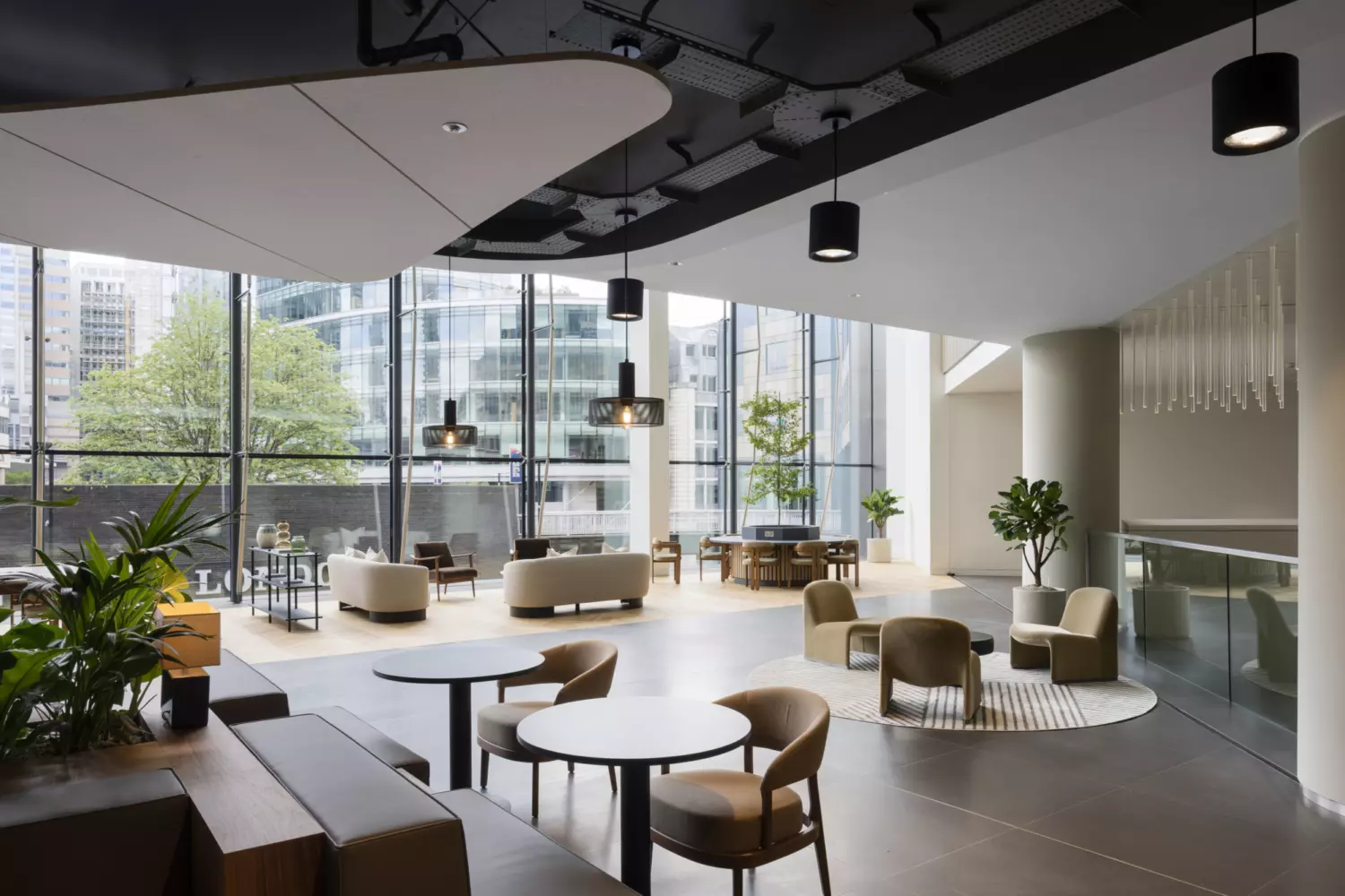 A spacious modern lounge area with floor-to-ceiling windows offering city views. Seating includes round tables with chairs, sofas and armchairs arranged for comfort and flexibility. Hanging light fixtures and ceiling panels add visual interest, while potted plants bring greenery to the contemporary, welcoming interior.