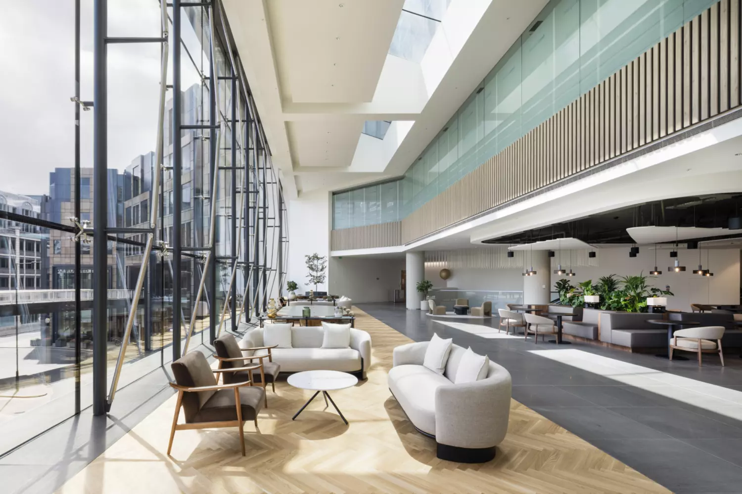 A spacious modern office interior with floor-to-ceiling windows on the left, flooding the space with natural light. Contemporary seating includes sofas and armchairs around small tables. Light wood and dark tile flooring define zones, while pendant lights, plants and glass panels with wooden slats enhance the sleek, welcoming design.