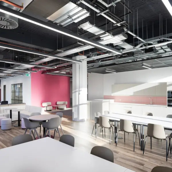 Modern open plan office breakout and kitchen area with a clean, contemporary fit out. The space features long white dining tables with light grey chairs in the foreground, alongside smaller round tables and soft seating to the left. At the back right, a white kitchenette includes integrated appliances, cabinets and a pink tiled splashback. A bold pink feature wall sits behind two white lounge chairs, adding contrast to the otherwise neutral palette. The ceiling is exposed, showing ventilation, cable trays and linear LED lighting, while large windows and overhead lights create a bright, evenly lit environment. The flooring is a mix of wood effect vinyl and darker carpet in the seating area.
