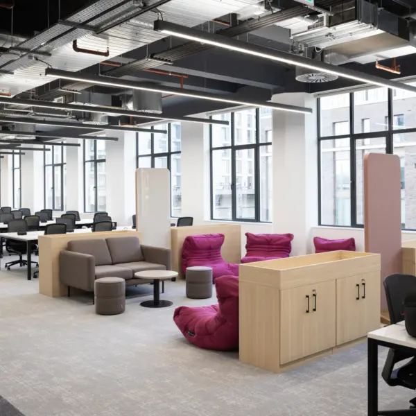 Modern open plan office breakout and workspace area with a contemporary fit out and exposed ceiling showing ductwork, cable trays and linear LED lighting. Large floor‑to‑ceiling windows line the back wall, bringing in natural light and views of neighbouring buildings.  In the foreground, a soft seating area features a light grey sofa, two round ottomans and a small circular table, positioned beside a cluster of bright pink lounge chairs with a relaxed, low profile design. Light wood storage units and integrated planters help define the seating zone and create separation from surrounding areas.  To the left, rows of white desks with black task chairs form a structured workspace, arranged in lines across the open floor. The flooring is a light grey finish throughout, keeping the palette neutral and cohesive, while the pink seating introduces a bold accent.  The space combines collaborative workstations with informal seating, offering a mix of focused work areas and relaxed breakout zones within a bright, clean and modern office environment.