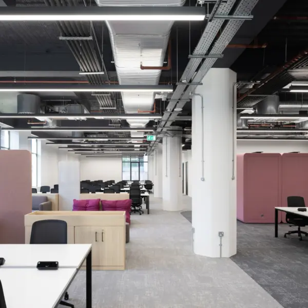 Modern open plan office workspace with a contemporary fit out and exposed ceiling featuring ductwork, cable trays and linear LED lighting arranged in parallel lines across the space. The layout is defined by large white structural columns that run through the centre, creating depth and subtle division between zones.  In the foreground, white desks with integrated power modules are paired with black task chairs, set on grey carpet tiles. To the left, a breakout seating area includes a light wood storage unit, a grey sofa and a cluster of bright pink lounge cushions positioned low to the floor, adding contrast to the neutral palette. A tall upholstered acoustic screen in soft pink stands nearby, helping to define the seating zone.  Further into the space, rows of desks extend towards the back, forming a structured working area with consistent black chairs and white surfaces. On the right, two large pink acoustic pods provide enclosed spaces for private calls or focused work, positioned near additional meeting tables and chairs.  Natural light enters from windows along the left side, while the combination of grey flooring, white walls and light wood finishes creates a clean, cohesive environment with bold pink accents throughout.