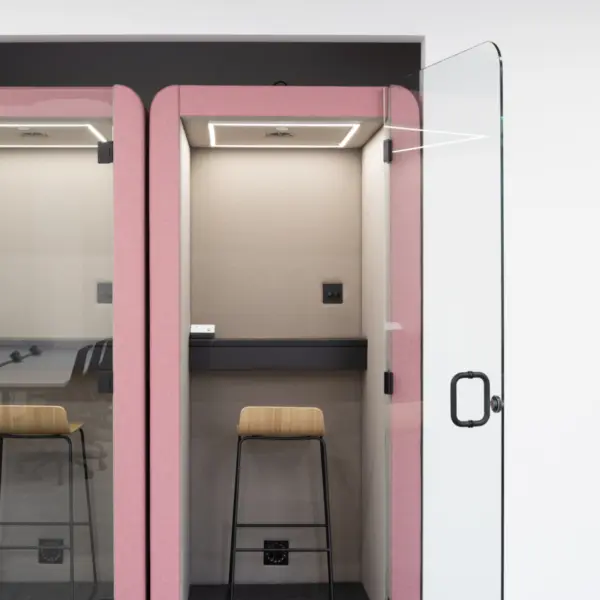 Modern office acoustic booths designed for private work or calls, finished in soft pink upholstered panels. Two adjoining pods are shown, with the right pod open and fitted with a clear glass door featuring a black handle.  Inside the open booth, a compact workspace includes a dark work surface, a high wooden stool with black legs and integrated power sockets on the back wall. A recessed ceiling light provides focused illumination within the booth, creating a quiet, enclosed environment.  The adjacent booth on the left is partially visible, also enclosed with glass and fitted with similar internal features. The surrounding space has a neutral finish with light grey flooring and white walls, allowing the pink booths to stand out as a bold yet functional element within the office fit out.