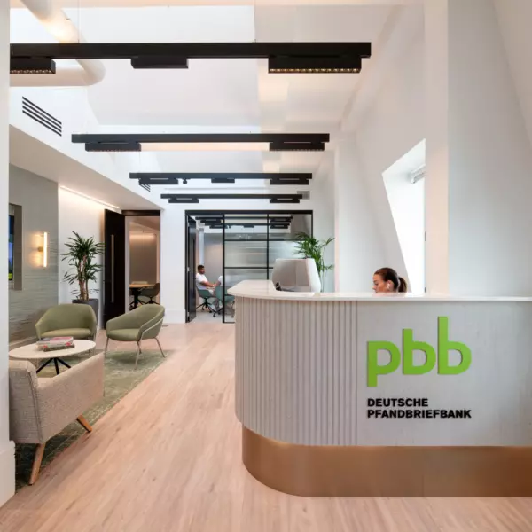 A contemporary office reception area at Deutsche Pfandbriefbank featuring a sleek white desk with the green pbb logo and company name. The space includes two seating zones with lime green chairs and minimalist tables. Natural light floods the room through large windows, complemented by recessed ceiling lights. Glass partitions reveal adjoining office areas, while strategically placed indoor plants add a touch of greenery and soften the modern aesthetic.