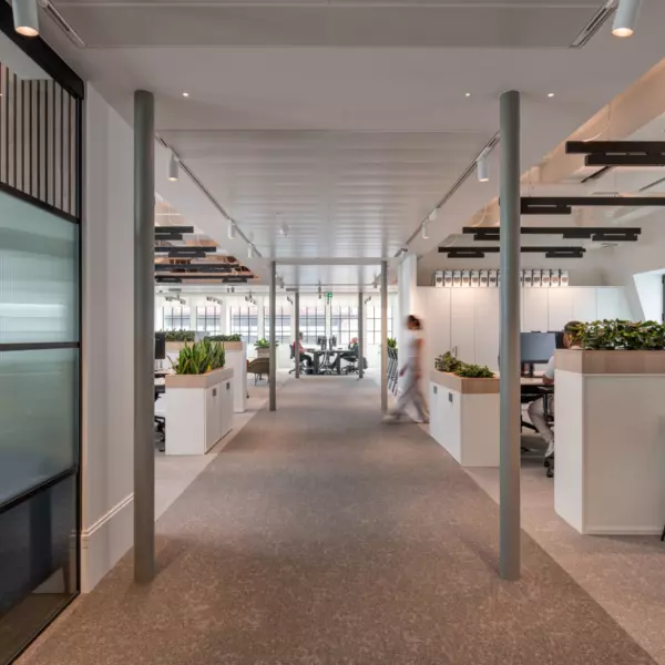 A modern open-plan office with symmetrical workstations flanking a central walkway lined with plants and low dividers. The ceiling features track lighting and structural columns. On the right, a high counter with green bar stools offers an informal workspace. The left side is defined by glass partitions with vertical detailing. Large windows in the background allow natural light to brighten the space, enhancing the clean, structured design.