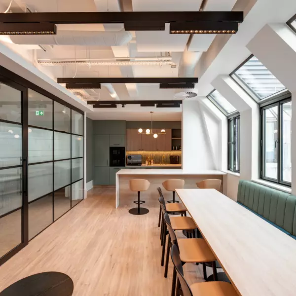 A modern office kitchen and dining area featuring a long wooden table with chairs on one side and a cushioned bench on the other. Large windows along the right wall flood the space with natural light. Glass partition walls on the left lead to another part of the office. The kitchen area in the background includes bar stools, pendant lights and built-in appliances such as a microwave and coffee machine. Exposed ceiling beams and sleek lighting fixtures complete the contemporary design.
