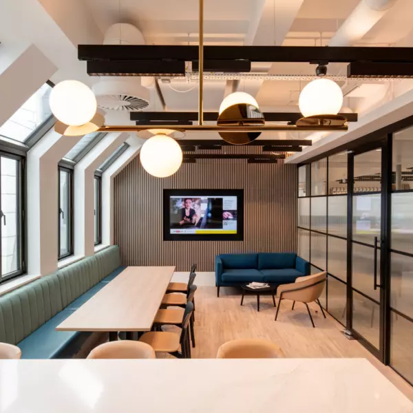 A stylish office lounge area featuring a long wooden table with chairs on the left and a seating zone with a blue sofa, matching armchairs and a wall-mounted television in the background. Large windows line one side of the room, allowing in natural light, while round pendant ceiling lights add a modern touch. Glass partition walls with vertical detailing define the right side, contributing to the open yet structured layout.