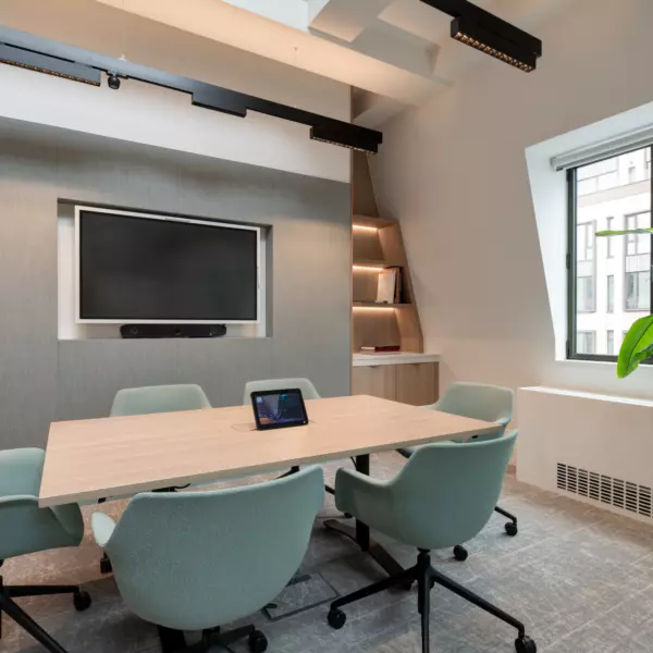 A contemporary conference room featuring a rectangular wooden table surrounded by five light green upholstered chairs on castors. A tablet rests on the table, while a wall-mounted flat-screen television with a soundbar sits opposite. Built-in shelves in the corner display books and decorative items. A large window brings in natural light, complemented by a tall potted plant nearby. The ceiling showcases exposed beams and track lighting, adding an industrial edge to the clean, modern design.