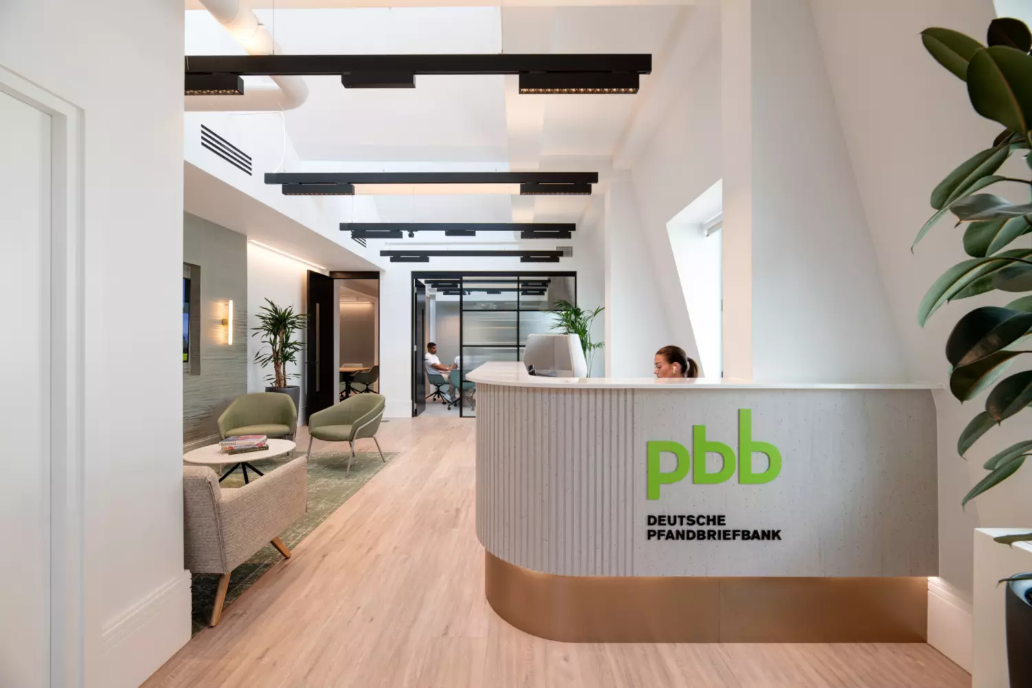 A contemporary office reception area at Deutsche Pfandbriefbank featuring a sleek white desk with the green pbb logo and company name. The space includes two seating zones with lime green chairs and minimalist tables. Natural light floods the room through large windows, complemented by recessed ceiling lights. Glass partitions reveal adjoining office areas, while strategically placed indoor plants add a touch of greenery and soften the modern aesthetic.