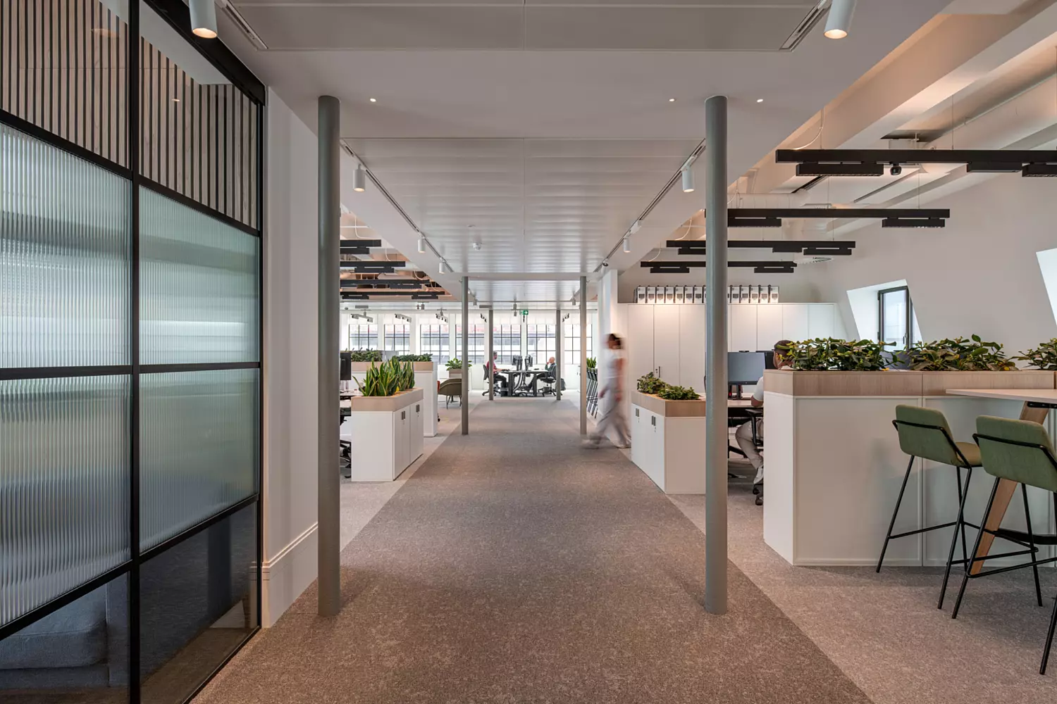 A modern open-plan office with symmetrical workstations flanking a central walkway lined with plants and low dividers. The ceiling features track lighting and structural columns. On the right, a high counter with green bar stools offers an informal workspace. The left side is defined by glass partitions with vertical detailing. Large windows in the background allow natural light to brighten the space, enhancing the clean, structured design.