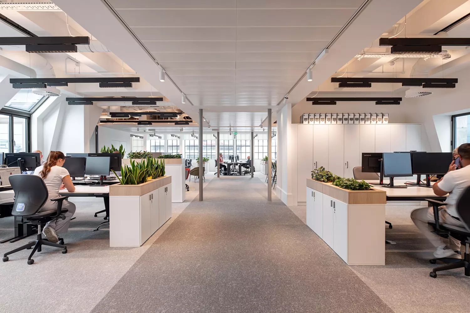 A modern open plan office featuring symmetrical rows of workstations flanking a central walkway. Each desk is equipped with a computer and an ergonomic chair. Large windows allow natural light to fill the space, supported by ceiling-mounted lighting. Low cabinets topped with green plants divide the walkway from the work areas, adding a natural touch to the clean, structured layout. The design promotes functionality and a calm working environment.