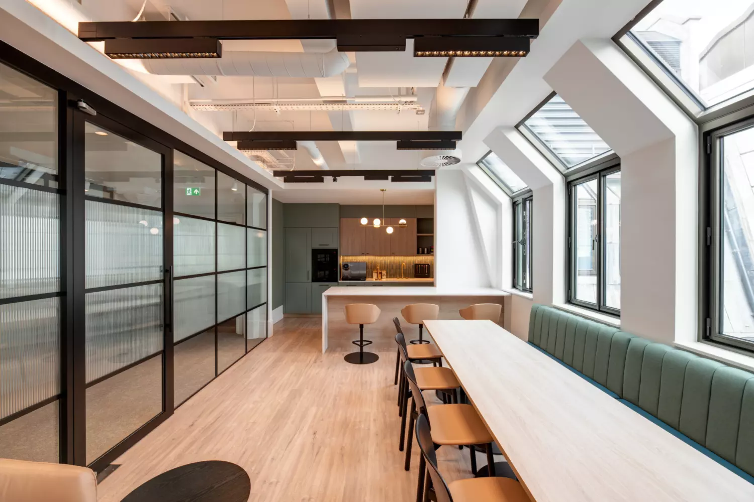A modern office kitchen and dining area featuring a long wooden table with chairs on one side and a cushioned bench on the other. Large windows along the right wall flood the space with natural light. Glass partition walls on the left lead to another part of the office. The kitchen area in the background includes bar stools, pendant lights and built-in appliances such as a microwave and coffee machine. Exposed ceiling beams and sleek lighting fixtures complete the contemporary design.