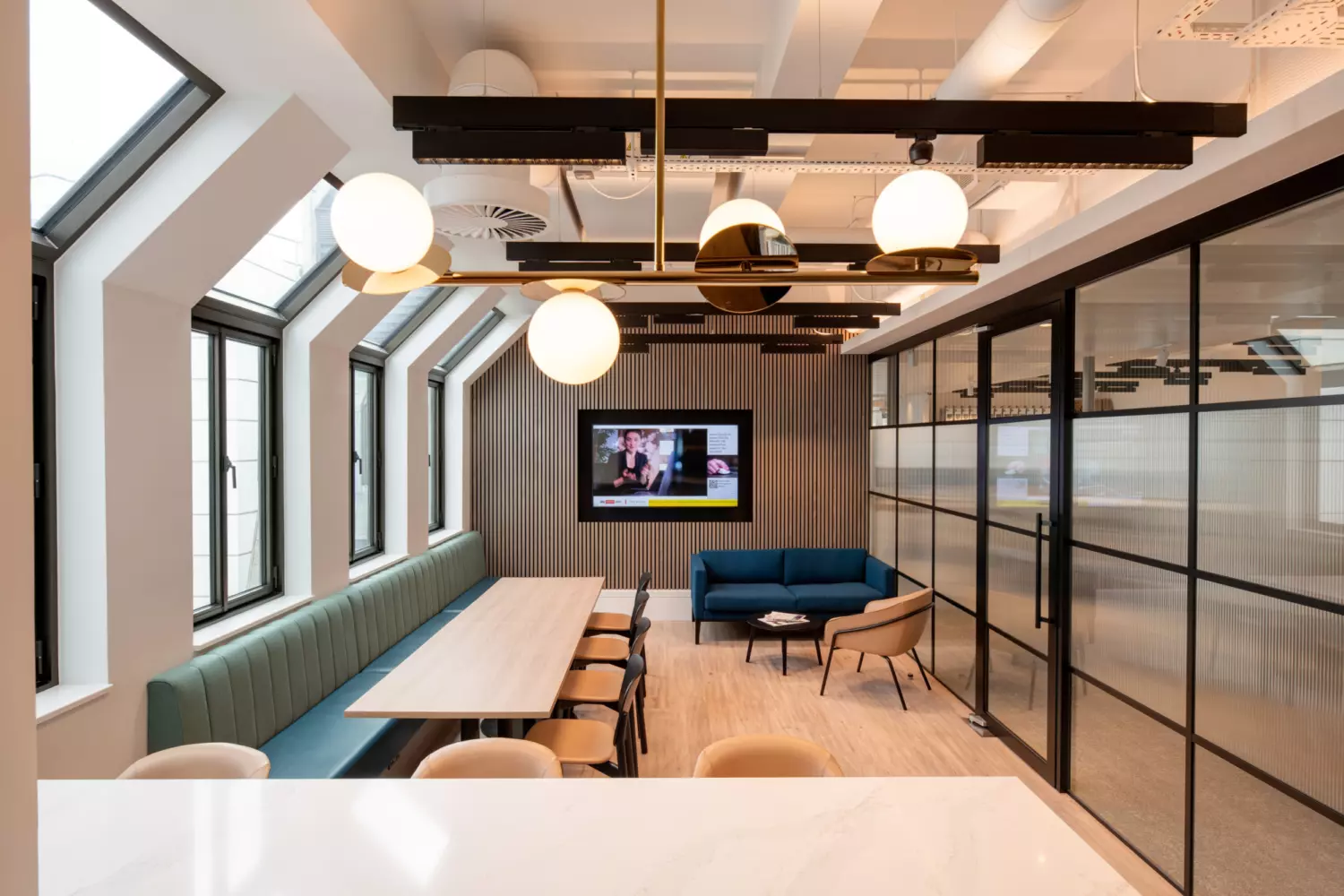 A stylish office lounge area featuring a long wooden table with chairs on the left and a seating zone with a blue sofa, matching armchairs and a wall-mounted television in the background. Large windows line one side of the room, allowing in natural light, while round pendant ceiling lights add a modern touch. Glass partition walls with vertical detailing define the right side, contributing to the open yet structured layout.