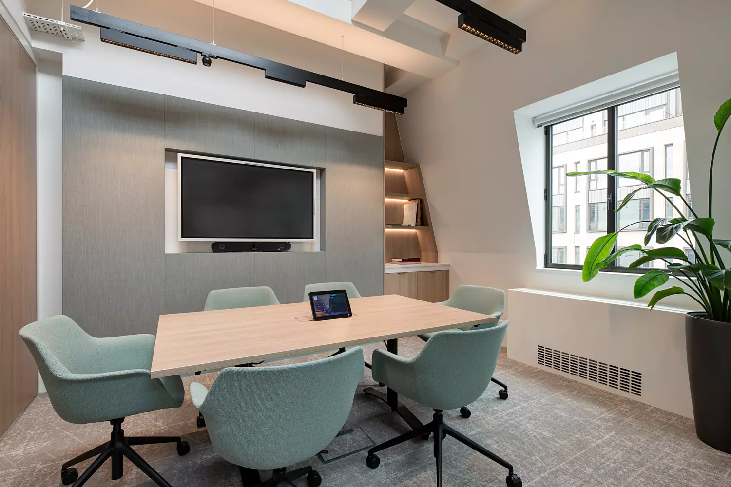 A contemporary conference room featuring a rectangular wooden table surrounded by five light green upholstered chairs on castors. A tablet rests on the table, while a wall-mounted flat-screen television with a soundbar sits opposite. Built-in shelves in the corner display books and decorative items. A large window brings in natural light, complemented by a tall potted plant nearby. The ceiling showcases exposed beams and track lighting, adding an industrial edge to the clean, modern design.