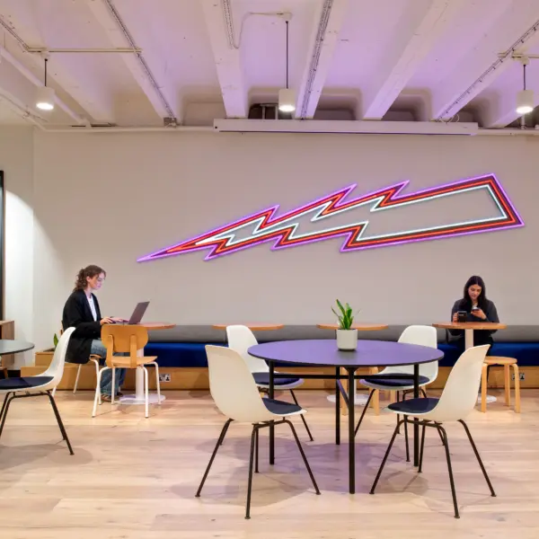 Large open-plan office breakout space with a mix of dining, collaboration and lounge areas. Round café-style tables and chairs are arranged on the left, while a long communal wooden table with woven-back chairs runs through the centre. A lounge seating area lines the right-hand wall beneath a black mountain-themed mural, and a neon wall feature provides a colourful focal point on the left. The space extends towards a kitchenette and a vibrant graffiti-style mural in the distance. Exposed ceilings, suspended lighting, glazed meeting rooms and indoor plants contribute to a bright, contemporary workplace environment.