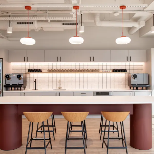 Contemporary office kitchenette featuring a large white island with three wooden bar stools and a deep red base. White cabinetry lines the back wall above a tiled splashback, with coffee machines, glassware and a sink arranged along the countertop. A drinks fridge stocked with canned beverages stands to the left, while floor-to-ceiling windows on the right bring natural light into the space. Exposed services, white-painted ceilings and pendant lights with red accents create a modern industrial-style aesthetic.