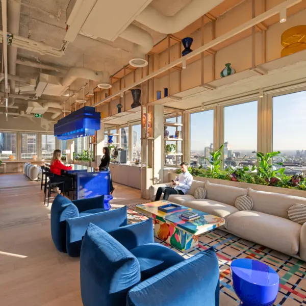 Modern office lounge with floor-to-ceiling windows overlooking the City of London skyline. The space features a large curved beige sofa, two blue lounge chairs, a colourful coffee table and abundant indoor plants. A glossy blue bar and matching suspended light fixture sit at the centre, while decorative glass vases line open shelving above. Three people occupy the space, with two seated at the bar and one seated on the sofa. Exposed ceilings, light wood finishes and natural daylight create a bright, contemporary workplace setting.