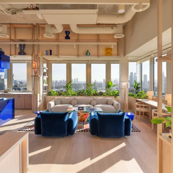 Contemporary office lounge with panoramic views across the London skyline through floor-to-ceiling windows. The space features a seating area with a curved beige sofa, four blue lounge chairs and a colourful coffee table set on a patterned rug. A glossy blue bar with matching pendant lighting sits to the left, while open timber shelving displays decorative glassware and integrated planters filled with greenery. Natural daylight floods the interior, complementing the light wood finishes, exposed ceiling services and vibrant design details throughout the workplace.