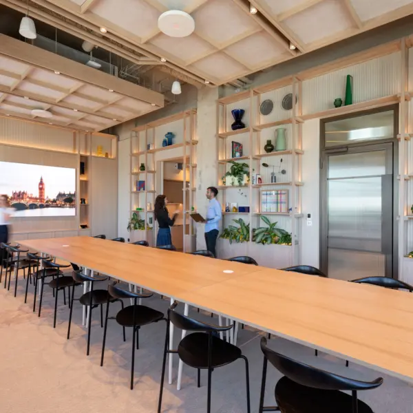 Contemporary meeting room with a long light wood conference table surrounded by black chairs. Floor-to-ceiling shelving lines the walls, displaying books, plants and colourful decorative glassware, with integrated lighting adding warmth throughout the space. A large screen at one end of the room shows a cityscape image, while exposed services and a coffered ceiling create a blend of modern and industrial design. Three people are visible within the room, with two standing in conversation and one walking past the table.