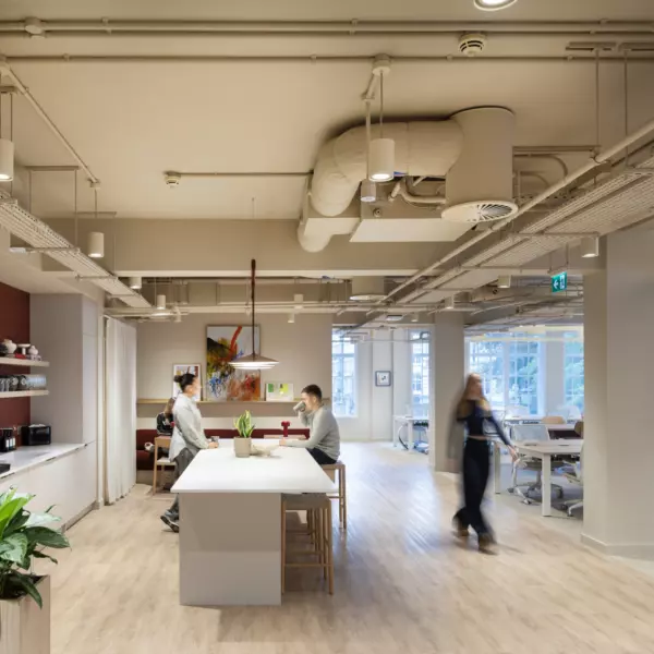 Modern open plan office with a bright, minimalist design featuring a central communal table with high stools, integrated coffee station and natural wood flooring. Exposed ceiling services and suspended lighting create an industrial aesthetic, complemented by large windows for natural light and greenery for a biophilic touch. Flexible workstations are visible in the background, promoting collaboration and contemporary workplace design