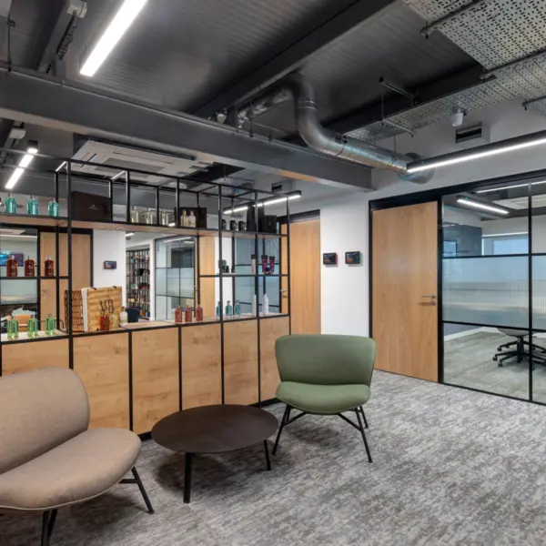 A contemporary office lounge area with two low armchairs, one in beige and one in green, arranged around a small circular coffee table on a grey patterned carpet. Behind them, a floor to ceiling shelving unit with black framing and wood panels displays assorted bottles and decorative items. Four timber doors line the surrounding white walls, and the ceiling features exposed ductwork, cable trays and linear LED lighting.