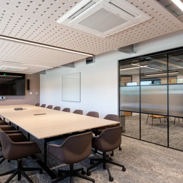 A modern meeting room with a large rectangular light wood table surrounded by swivel chairs upholstered in dark brown fabric. A wall mounted screen and video conferencing unit sit at the far end of the room above a wood credenza. The space has a grey patterned carpet and a ceiling with exposed services and perforated acoustic panels. To the right, a glass partition with black framing and fluted panels separates the room from an adjacent workspace, with a timber door set into the partition.