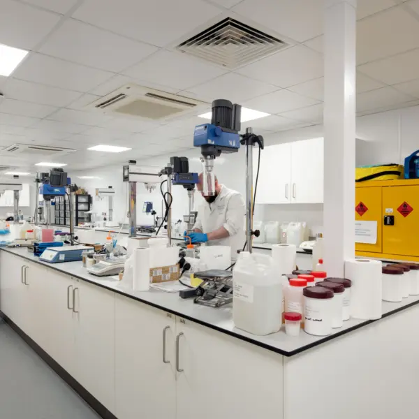 A bright laboratory with white walls, ceiling panels and long central workbenches. Containers, instruments and mixing equipment are arranged across the surfaces. Two people in lab coats and gloves are working at separate stations, one using a bench mixer and the other preparing materials. Safety cabinets in bright yellow stand against the right wall, and various pieces of technical equipment and storage line the background.