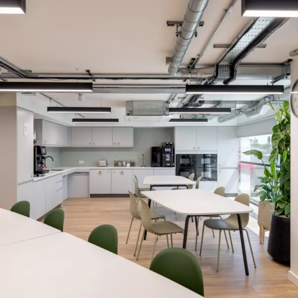 A bright tea point and breakout area with white worktops, pale green chairs and light wood flooring. The kitchen includes white cabinets, a coffee machine and small appliances set against a tiled splashback. Overhead, exposed ductwork and linear lights run across the ceiling. Large windows bring in natural light, and a tall potted plant sits to the right beside a wall clock.
