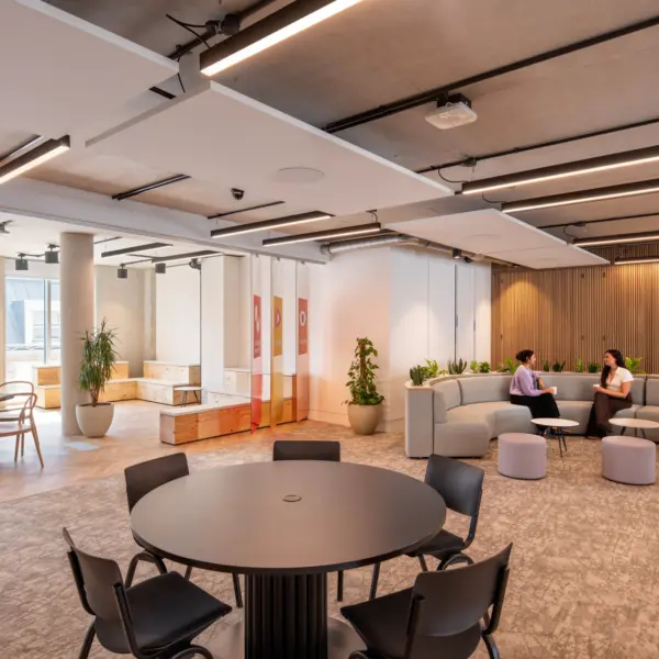 Open-plan office with a round meeting table and chairs in the foreground. A curved grey sofa and small stools form an informal seating area, where two people are seated facing each other. Large windows, exposed concrete columns, indoor planting and a timber slatted wall add warmth to the space, while linear ceiling lights run across the exposed concrete ceiling.