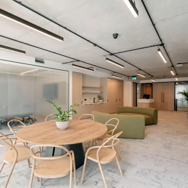 Open office breakout space with a large round timber table and light wooden chairs in the foreground. Green upholstered seating sits beyond, alongside pale timber storage, shelving and a reception desk. A glass-fronted meeting room labelled “Sudbrook” is on the left. Exposed concrete ceilings, linear lights, timber finishes, indoor planting and neutral carpet complete the space.