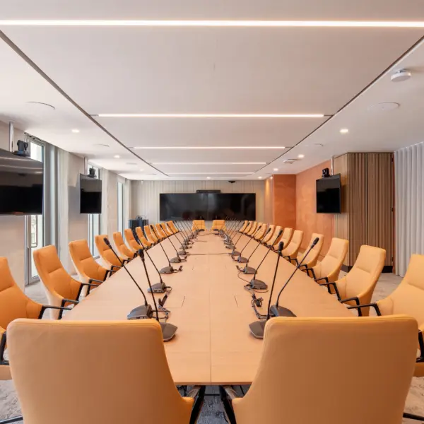 Large boardroom with a long U-shaped timber conference table surrounded by orange chairs. Individual microphones are positioned around the table, with several wall-mounted screens and cameras for video conferencing. Floor-to-ceiling windows run along one side, while timber panelling and white curtains line the opposite walls. Linear ceiling lights illuminate the room.