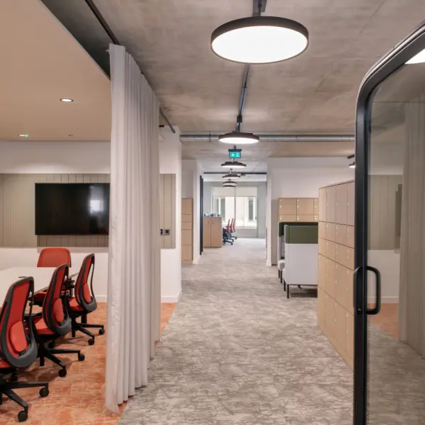 Modern office corridor with an exposed concrete ceiling and circular pendant lights. A meeting area with a long table, red office chairs, wall-mounted screen and timber slatted wall sits to the left, separated from the corridor by a curtain. Lockers and work booths line the right side, with a dark framed glass meeting pod in the foreground.