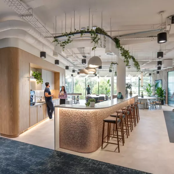 A contemporary office interior showcasing a central curved bar counter with high stools, lit by pendant lights. The bar features a textured façade and is accentuated by ceiling-mounted greenery. To the left, a wooden kitchenette includes a coffee machine and informal gathering space. On the right, semi-private booth seating with frosted glass panels offers quieter work zones. The flooring combines light tiles with dark carpet, while expansive windows provide ample natural light. Exposed ductwork and suspended lighting contribute to the open-plan, industrial aesthetic.