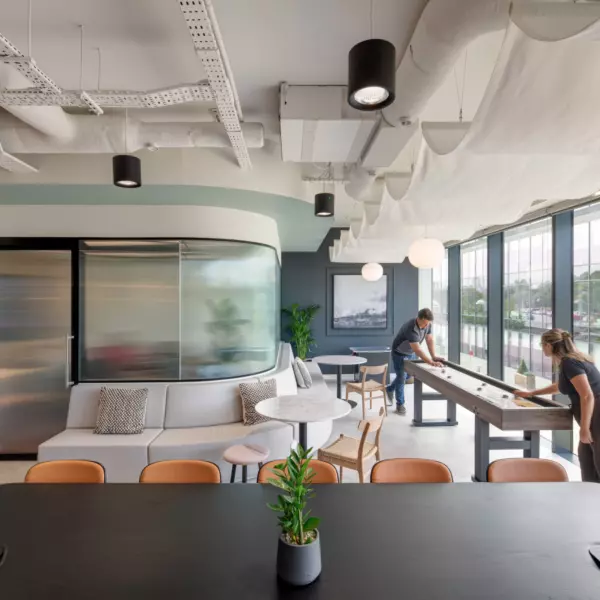 A stylish office lounge area with large windows providing ample natural light. The space includes a curved sofa, chairs and stools arranged around tables offering varied seating options. A shuffleboard table is positioned on the right adding a recreational element. The ceiling features exposed ductwork and suspended lighting contributing to the industrial aesthetic. Indoor plants placed on tables and near the back wall enhance the relaxed modern atmosphere.