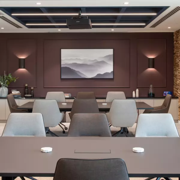A modern conference room with a large central table surrounded by multiple chairs. A ceiling-mounted projector faces a wall-mounted screen displaying an abstract mountain image. Another screen on the right wall shows additional content. The room features wooden wall panelling with shelves holding plants and decorative items, creating a warm professional atmosphere.