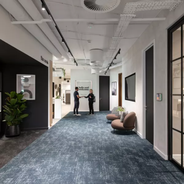 A modern office hallway with blue carpet and white walls. Two people are standing in the centre of the corridor having a conversation. The ceiling features exposed pipes and ducts with multiple light fixtures. On the left, a black-framed glass door leads to another room with a large potted plant beside it. On the right, two brown chairs with pillows are placed against the wall near another black-framed glass door.