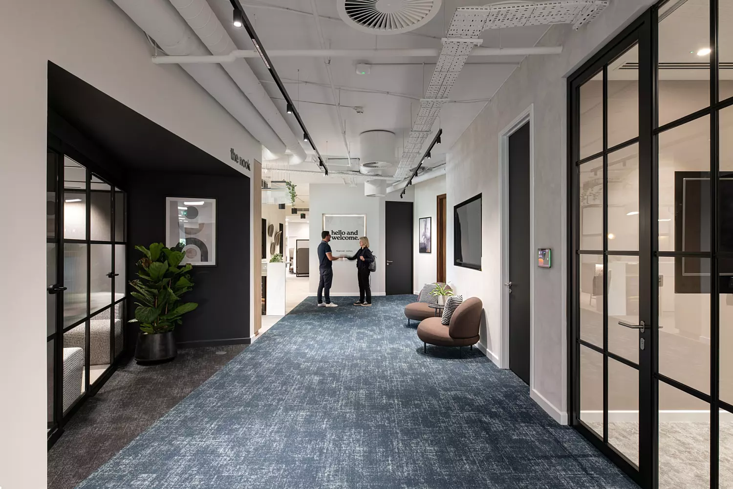 A modern office hallway with blue carpet and white walls. Two people are standing in the centre of the corridor having a conversation. The ceiling features exposed pipes and ducts with multiple light fixtures. On the left, a black-framed glass door leads to another room with a large potted plant beside it. On the right, two brown chairs with pillows are placed against the wall near another black-framed glass door.