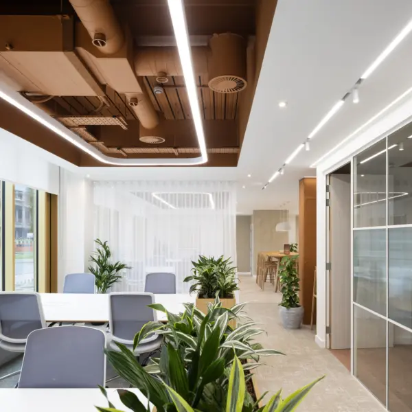 Modern office interior with a clean, minimalist design. In the foreground, a white meeting table is surrounded by several light grey office chairs, with multiple green potted plants placed around the seating area. Large windows on the left let in natural light and show a street scene outside. The ceiling features exposed ductwork painted in a warm brown tone, contrasted with linear LED light strips. In the background, sheer white curtains partially divide the space, leading to a casual breakout area with high wooden stools and a counter. On the right side, a glass-partitioned room with a sliding door is visible. The overall space uses a mix of neutral colours, warm wood accents and indoor greenery to create a bright, contemporary workplace.