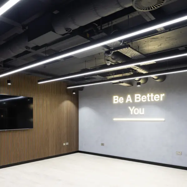 A contemporary office feature wall within a modern interior space. The main wall is finished in a smooth, light grey surface, displaying the illuminated text “Be A Better You” in soft white lettering, with a thin light bar beneath it. To the left, a large wall-mounted black screen sits against a contrasting vertical slatted wood panel.  Above, the ceiling is open, revealing exposed ductwork, cables and services painted in dark tones, paired with several long, linear LED light fittings that run parallel across the space. The floor is a light wood-effect finish, keeping the area bright and uncluttered. The overall scene has a clean, minimal look, combining industrial ceiling elements with warm wood and soft, indirect lighting to create a bold focal point within the office.