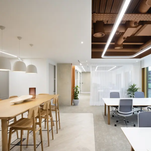 Wide view of a modern office interior that blends a breakout area with open workspaces. On the left, a long light wood high table is surrounded by matching wooden stools, with three large white pendant lights hanging above to create a soft, focused glow. The table sits on a light wood-effect floor, which transitions into a carpeted area on the right.  In the centre background, a sheer white curtain hangs from ceiling to floor, partially dividing spaces while still allowing light to pass through. Behind the curtain, additional seating and lighting are faintly visible. A corridor with neutral-toned walls and a door extends beyond this area, adding depth to the layout.  On the right, several white desks with grey office chairs are arranged near large windows that bring in natural daylight and offer a view of a bus and street outside. A potted green plant is placed near the window, adding a natural element.  Above, the ceiling combines a clean white finish with a feature section of exposed services painted in a warm brown tone, framed by continuous LED strip lighting. The overall palette of soft whites, warm wood and greenery creates a bright, calm and contemporary workplace environment.