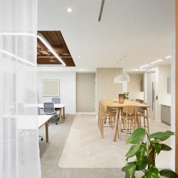 Wide view of a contemporary office interior combining work and breakout spaces. In the foreground on the right, a green potted plant sits beside a textured terracotta-coloured wall. To the left, sheer white floor-to-ceiling curtains partially divide the space, softly diffusing light. Behind the curtains, a work area with white desks and grey office chairs is visible, set on a darker carpeted floor.  In the centre-right, a light wood high table with several matching stools forms a casual meeting or social area. Above the table, three white pendant lights hang in a row, while recessed ceiling lights and linear LED strips provide additional illumination. The ceiling includes a feature section with exposed services painted in a warm brown tone, framed by integrated lighting.  Further back, neutral-toned walls and cabinetry line the corridor, with a door and small kitchenette-style units visible. The overall space uses a palette of white, beige, warm wood and terracotta accents, complemented by indoor plants and soft lighting to create a bright, modern workplace.