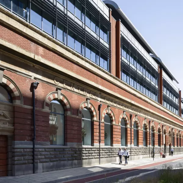 A long multi storey building with a red brick facade and large windows. The lower level features arched windows framed by decorative stonework. The words "Central Electric Lighting Station" are displayed above the arches. People walk along the pavement in front, adding scale to the historic yet functional design.