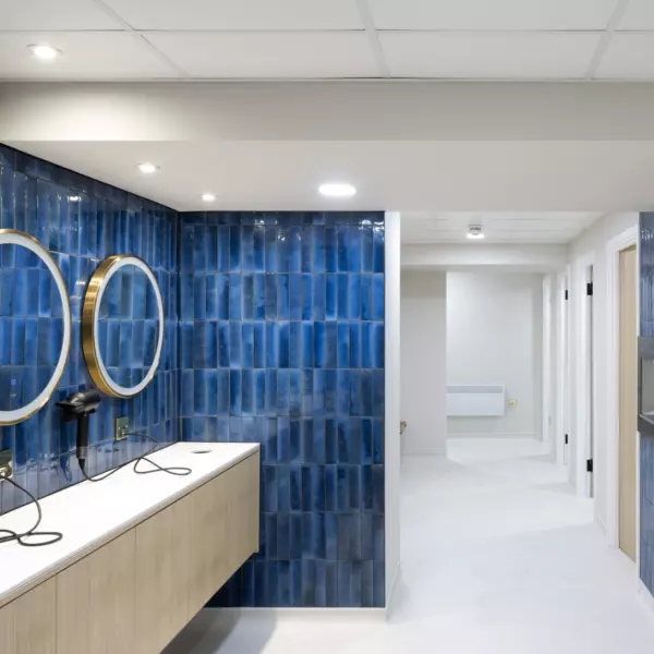 A modern changing area with blue tiled walls, round gold framed mirrors and wall mounted hairdryers. A hallway leads to several doors, likely for individual stalls or rooms. Recessed ceiling lights and light coloured tiled flooring contribute to the clean, functional design.