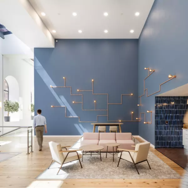 A modern interior space with high ceilings and large arched windows that fill the room with natural light. The seating area features pink and beige furniture on a light rug, set against a blue wall with geometric light fixtures. Potted plants and soft colours create a relaxed, contemporary atmosphere.