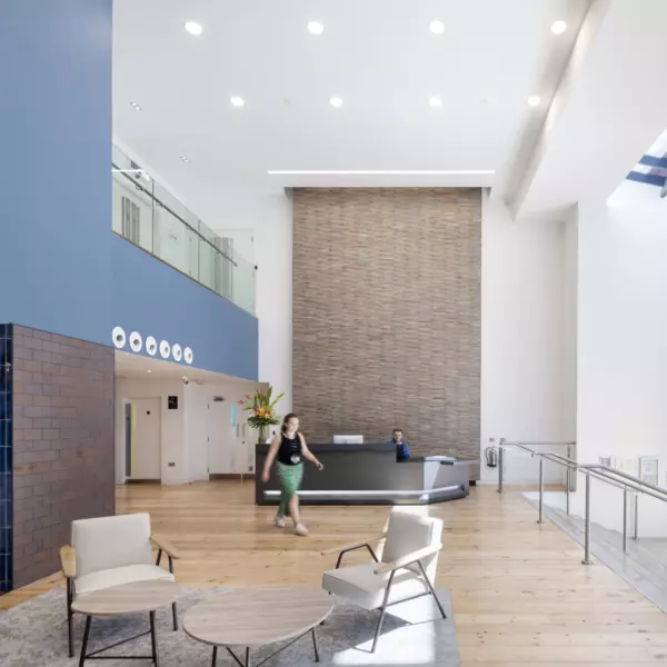 A spacious modern lobby with high ceilings and natural light streaming through large windows. The reception desk sits against a tall brick wall, while the seating area includes two chairs and round tables on a rug. Blue tiled walls and wooden flooring add texture and warmth to the contemporary design.