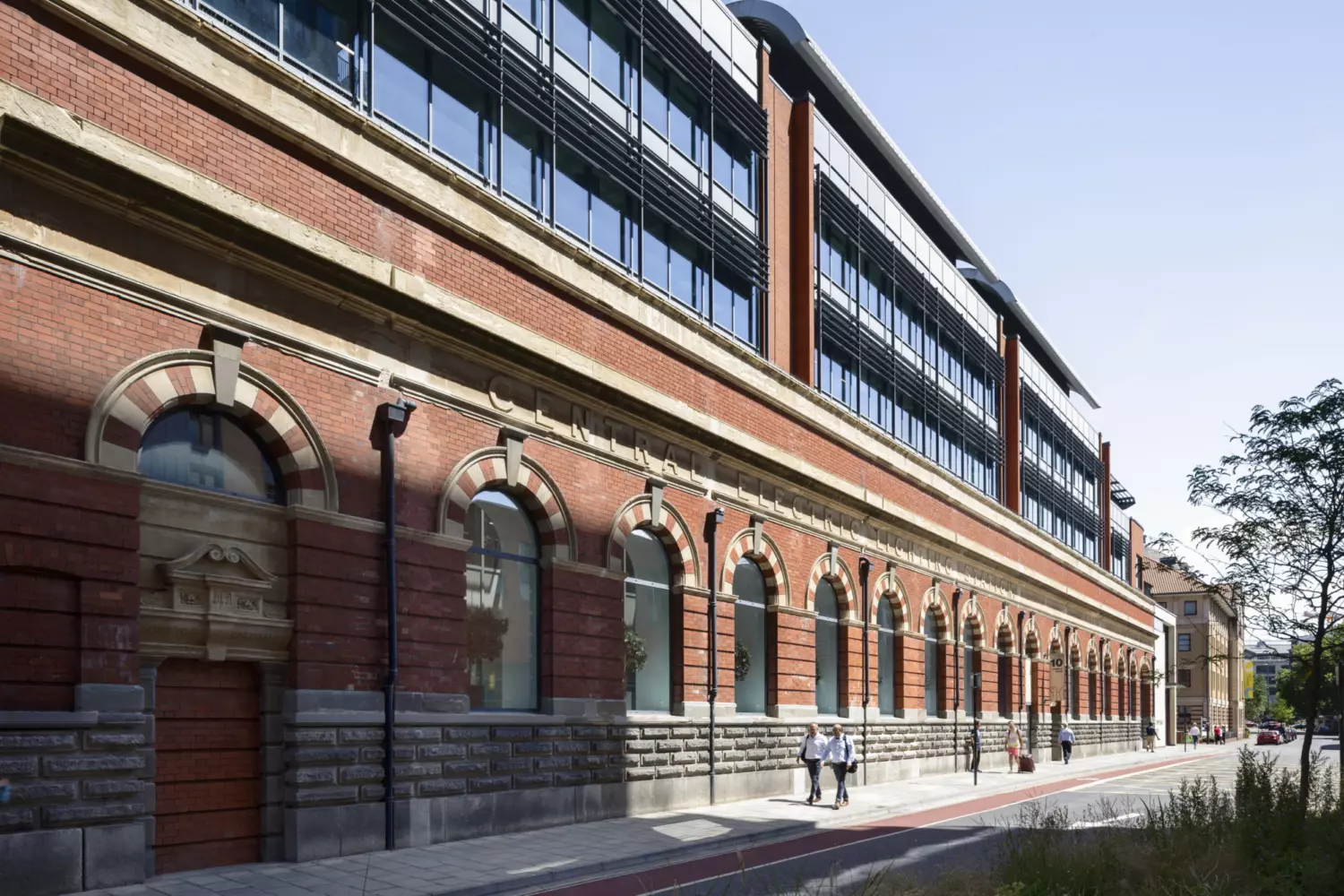 A long multi storey building with a red brick facade and large windows. The lower level features arched windows framed by decorative stonework. The words "Central Electric Lighting Station" are displayed above the arches. People walk along the pavement in front, adding scale to the historic yet functional design.
