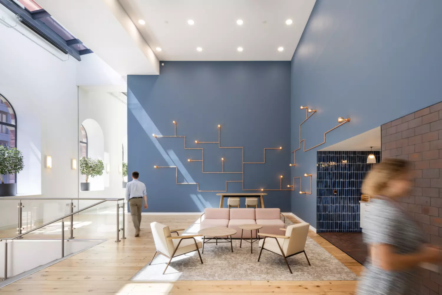 A modern interior space with high ceilings and large arched windows that fill the room with natural light. The seating area features pink and beige furniture on a light rug, set against a blue wall with geometric light fixtures. Potted plants and soft colours create a relaxed, contemporary atmosphere.